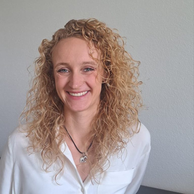 Eine lächelnde Frau mit lockigem, blondem Haar und einem weißen Oberteil mit schwarzem Muster.